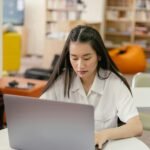 A young woman uses a laptop in a cozy library setting, emphasizing education and technology.